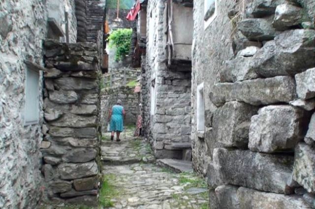 Senhora caminha por uma rua estreita de Corippo, ladeada por construções de pedra (Foto: BBC) Senhora caminha por uma rua estreita de Corippo, ladeada por construções de pedra (Foto: BBC)
